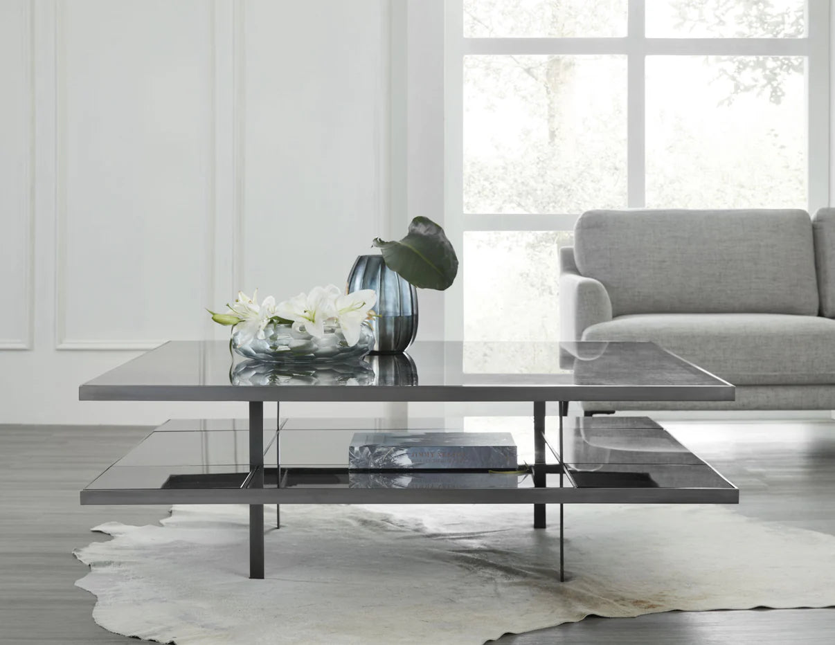 In this lifestyle photo, The Romo Cocktail Table is seen in an oil-rubbed bronze finish. It's a perfect blend of modern design and timeless elegance, making it an ideal centerpiece for any contemporary living space.
Available in two sizes—54" x 54" x 18" and 60" x 60" x 18"—this square cocktail table features a sleek metal base paired with a clear glass top that exudes sophistication and style. Its minimalist lines and balanced proportions make it a versatile choice for both large and small living rooms.