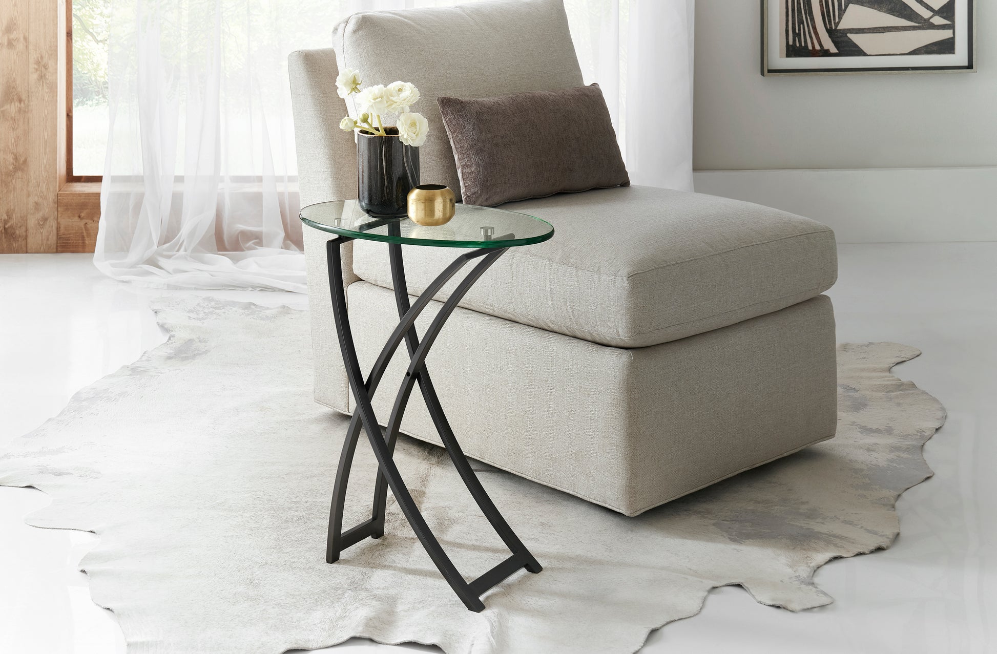Pictured in this lifestyle photo in an oil-rubbed bronze finish, The Playa Side Table is an elegant blend of modern design and artistic sophistication, perfect for adding a touch of contemporary style to any room.
This metal and glass end table features an oval-shaped glass top that sits gracefully on a metal frame with subtle, flowing curves. Its sleek oil-rubbed bronze finish enhances the table's refined look, making it an ideal choice for living rooms, bedrooms, or lounge areas.