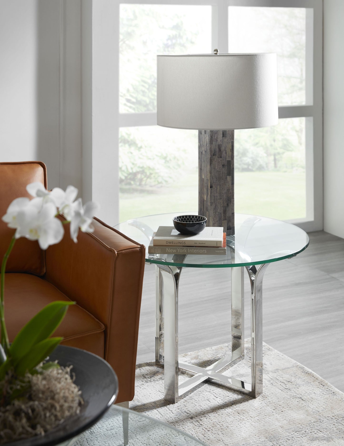 Pictured here in this lifestyle shot, The Trellis Round Accent Table features a sleek stainless steel base inspired by the intricate geometric patterns of a traditional trellis design, this round accent table offers a visually captivating element to your space.
Topped with a clear glass tabletop, it provides a light, airy feel that complements a variety of interior styles.