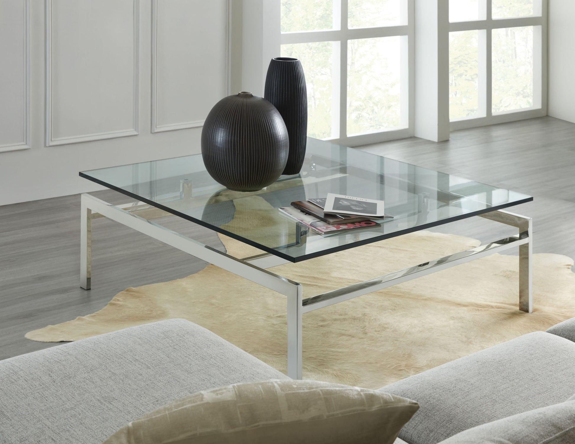 In this lifestyle photo, we see The Lance Cocktail Table, a quintessentially modern piece that encompesses simplicity and timeless elegance. This square cocktail table features a sleek stainless steel frame paired with a clear glass top, creating a clean and contemporary look that complements any decor style.