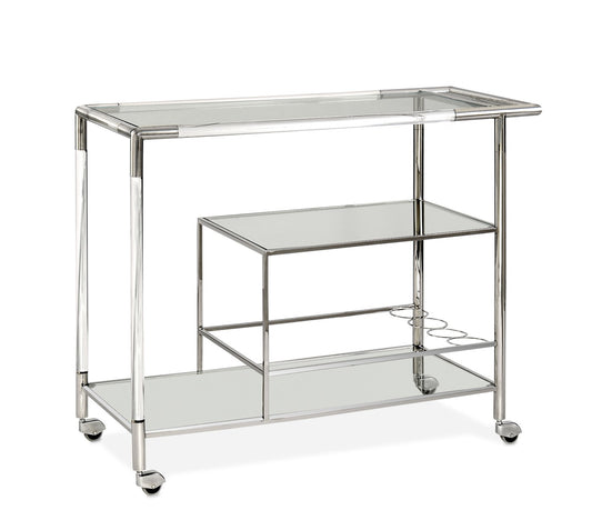 Pictured here, the Lumen Bar cart is crafted from polished stainless steel. This mobile bar cart features a spacious glass top shelf and a smaller nesting glass shelf below, offering ample space for displaying bottles, glassware, and bar accessories. Whether used as a stationary serving bar or a dynamic, mobile solution for parties and events, the Lumen Bar Cart effortlessly elevates any occasion.