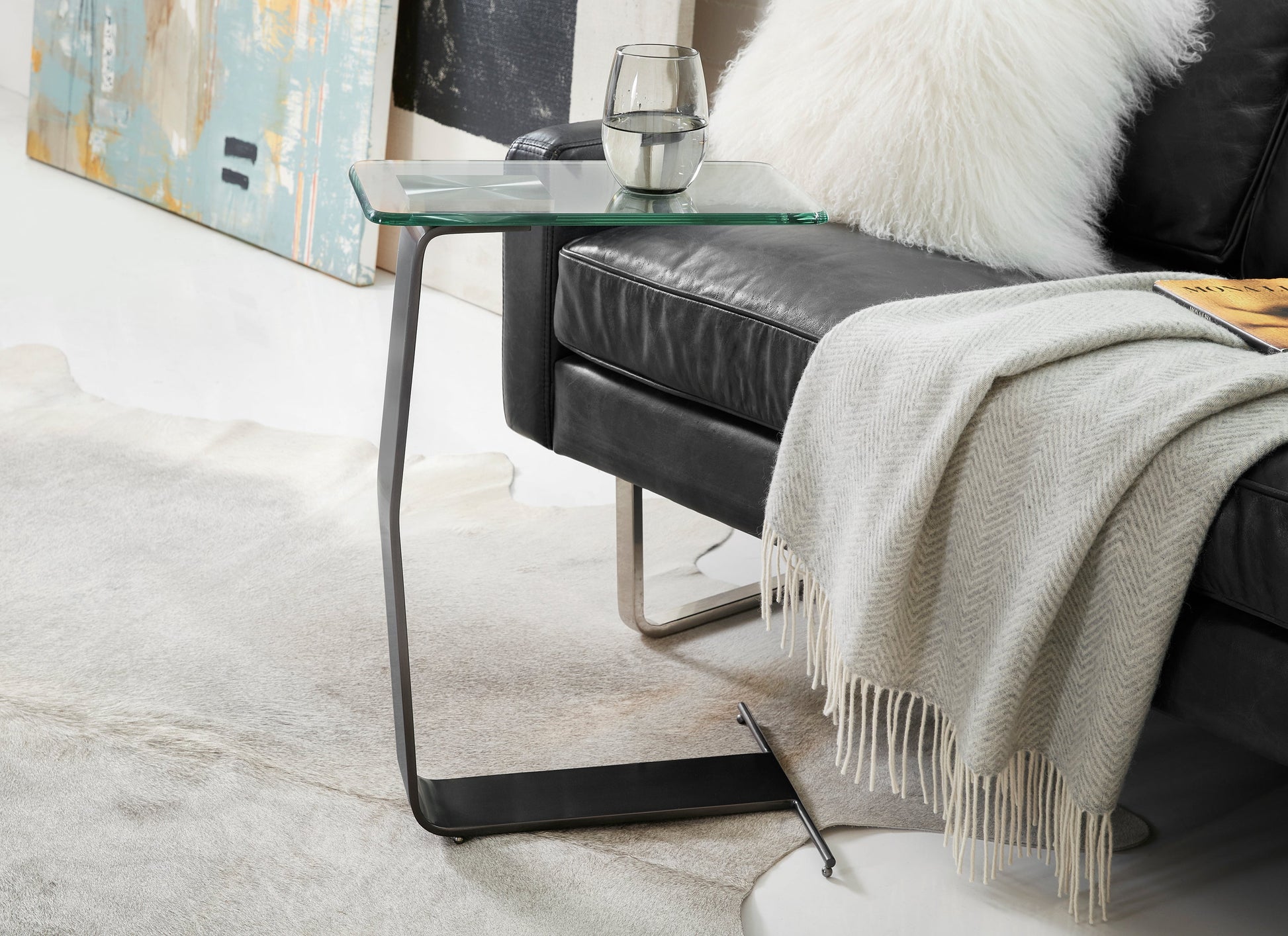 This is a lifestyle photo of the Plateau Side table shown in oil-rubbed bronze.

Featuring a clear glass tabletop that rests on a sleek, sculptural metal frame, this metal and glass side table adds a touch of sophistication to your home decor. The unique frame, crafted from a single piece of metal, creates a contemporary sculpture-like form that serves as both a functional end table and a stylish accent piece.