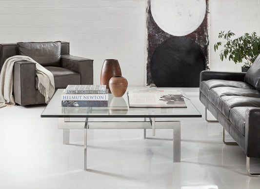 Pictured in this lifestyle shot, The Zig Zag Square Cocktail Table is a striking addition to any modern living room or lounge space, offering both functionality and artistic flair.

Crafted with a sleek stainless steel frame, this square cocktail table features an intricate base design that creates an eye-catching optical illusion. The geometric legs appear to twist and intersect beneath the clear glass tabletop, adding a unique architectural element to your decor.