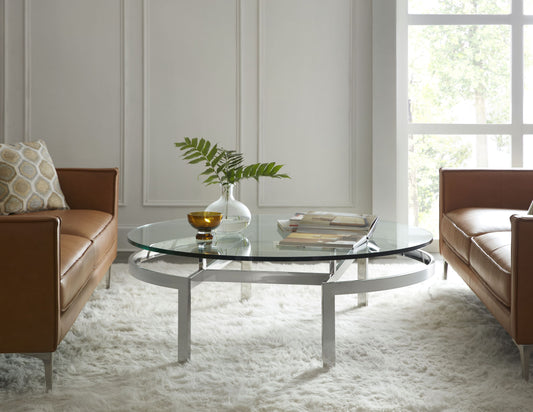 Pictured in this lifestyle shot is the Zig Zag Round Cocktail table by Modern Elements furniture. Featuring a stainless steel frame with an intricate base design, this round cocktail table creates an optical illusion with its legs, appearing to twist and turn as they connect seamlessly to the clear glass tabletop.